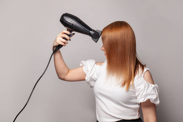 Όμορφη νεαρή γυναίκα στεγνώνει τα κόκκινα μαλλιά της με στεγνωτήριο πάνω από γκρι φόντο - Φωτογραφία, εικόνα