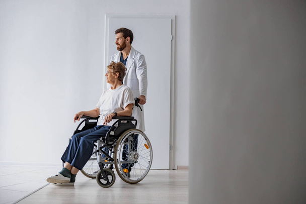 Klinikte tekerlekli sandalyeli hasta taşıyan profesyonel bir erkek doktor. Yüksek kalite fotoğraf - Fotoğraf, Görsel