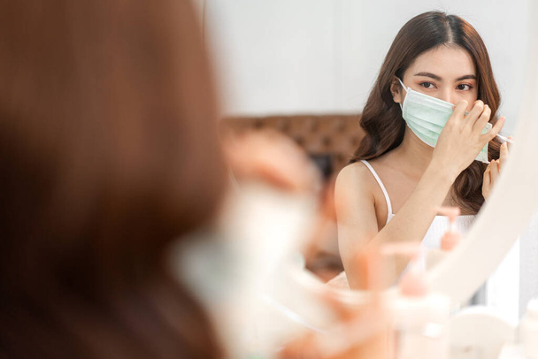 Young asian woman clean fresh healthy white skin looking at mirror in quarantine for coronavirus wearing surgical mask face protection with social distancing at home.covid19 and new normal concept - Foto, afbeelding