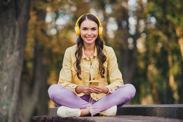 Retrato de bela menina alegre na moda sentado no banco usando dispositivo gadget ouvindo melodia alma descansando ao ar livre. - Foto, Imagem