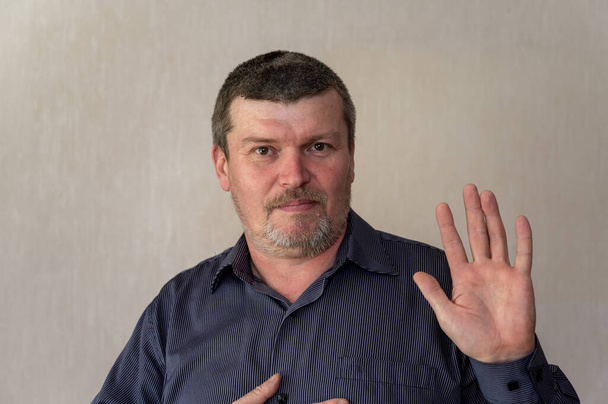 Portret van een volwassen man die een handgebaar van begroeting maakt. Een man in een blauw gestreept shirt zwaait met zijn hand in de lucht. Kort bruin haar met grijs. In de kamer. Selectieve focus. - Foto, afbeelding