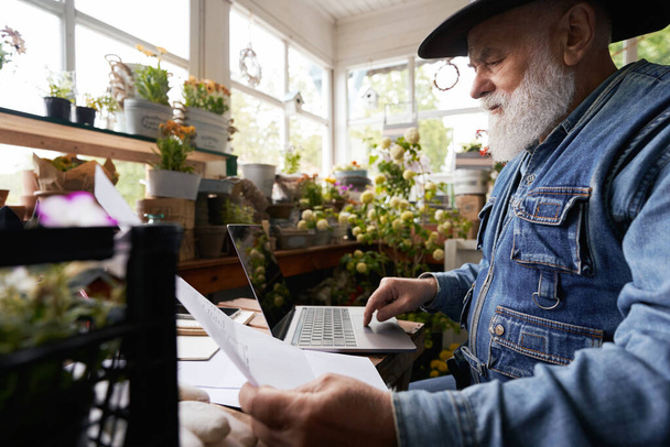 帽子のシニア男性はノートに入力し、開花ポット植物の間で机に座っている間紙を保持しています - 写真・画像