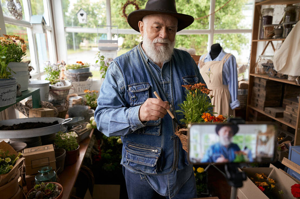 Uomo con la barba d'argento sta mostrando come prendersi cura di piante in vaso online e utilizzando la fotocamera - Foto, immagini