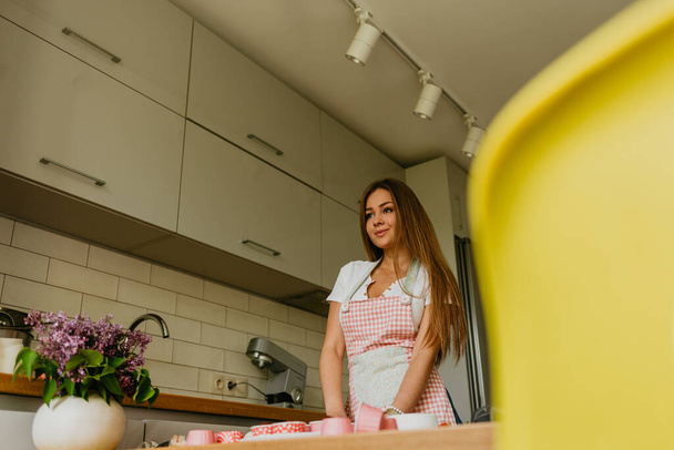 Portrait of friendly smiling female professional confectioner topping a cupcake with cream using a pastry bag. Looking at the camera. Indoors image. Pastry chef woman making creamy cakes - Foto, Imagem