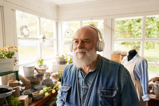 Joyeux homme âgé avec barbe argentée utilise des écouteurs pour s'amuser dans le magasin de fleurs - Photo, image