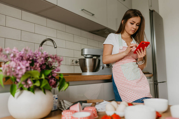 junge Frau mit Gemüse lächelt, während sie ihr Handy in der Küche benutzt - Foto, Bild