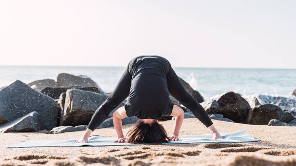 Takaisin näkymä tunnistamaton nainen activewear seisoo Prasarita Padottanasana matolla ja venyttely kehon aikana jooga istunnon hiekkainen meressä kesällä - Valokuva, kuva