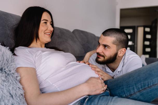 Porträt einer glücklichen Schwangeren mit Babyschuhen und ihres Mannes auf einem Sofa zu Hause - Foto, Bild