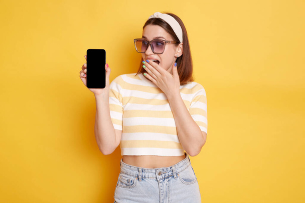Indoor shot of shocked surprised young woman wearing striped t-shirt posing isolated over yellow background, holding cell phone with empty display for advertisement. - Фото, изображение