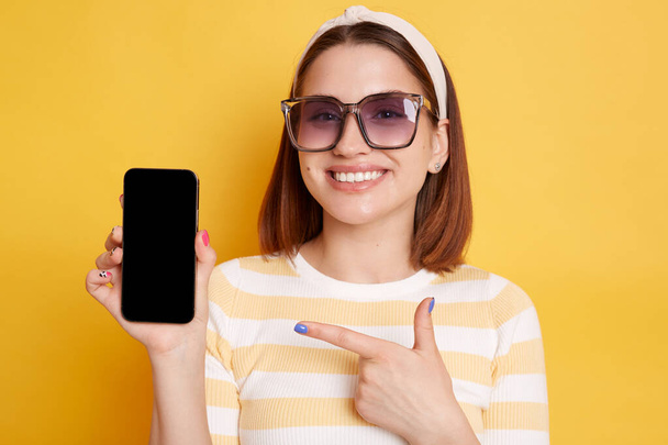 Horizontal shot of satisfied young woman wearing striped t-shirt posing isolated over yellow background, pointing at smart phone with blank screen for promotional text. - Foto, Imagem