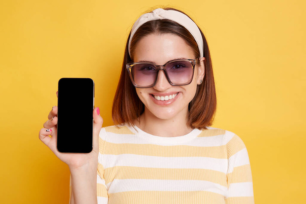 Indoor shot of delighted dark haired young woman wearing striped t-shirt posing isolated over yellow background, showing mobile phone with empty display for advertisement. - Foto, Imagem