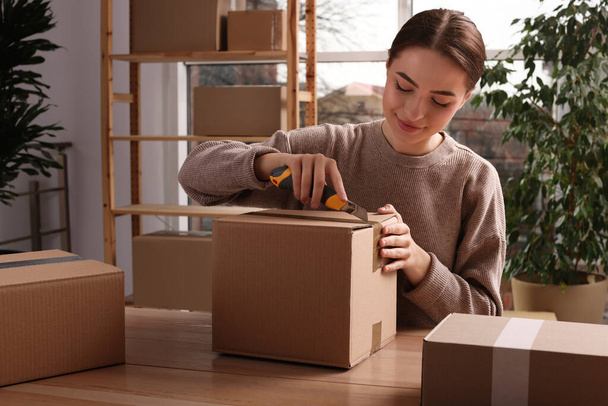 Young woman using utility knife to open parcel at wooden table indoors - Foto, imagen