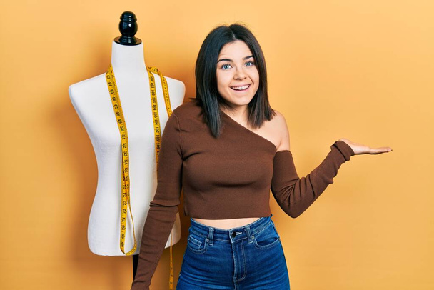 Young brunette woman standing by manikin celebrating achievement with happy smile and winner expression with raised hand  - Foto, afbeelding