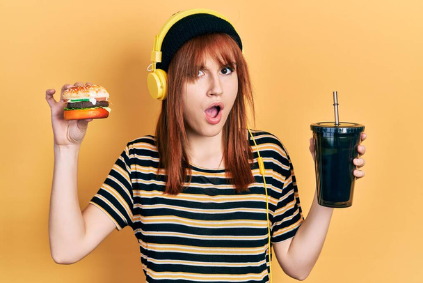 Rudowłosa młoda kobieta jedząca hamburgera i pijąca sodę ze słuchawkami w szokującej twarzy, wyglądająca sceptycznie i sarkastycznie, zaskoczona otwartymi ustami  - Zdjęcie, obraz