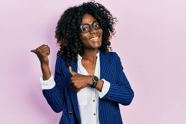Fiatal afro-amerikai nő, aki üzleti ruhát és szemüveget visel, feltartott kézzel és hüvelykujjal a háta mögé mutat, mosolyogva magabiztosan.  - Fotó, kép