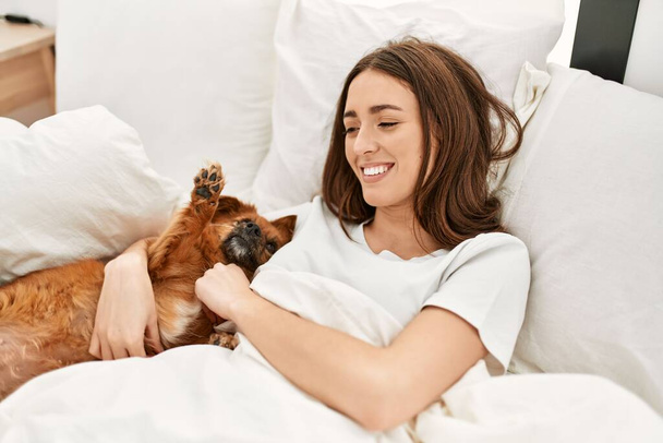 Joven mujer hispana abrazando perro acostado en la cama en el dormitorio - Foto, Imagen