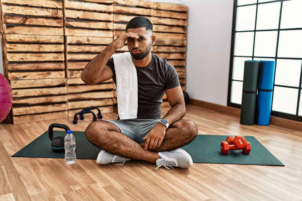 Νεαρός Ινδός που κάθεται στο πατάκι στο γυμναστήριο ανήσυχος και αγχωμένος για ένα πρόβλημα με το χέρι στο μέτωπο, νευρικός και ανήσυχος για την κρίση  - Φωτογραφία, εικόνα