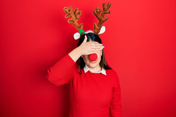 Mujer hispana joven con sombrero de Navidad de ciervo y nariz roja cubriendo los ojos con la mano, luciendo seria y triste. concepto de ocultamiento y rechazo sin visión  - Foto, imagen
