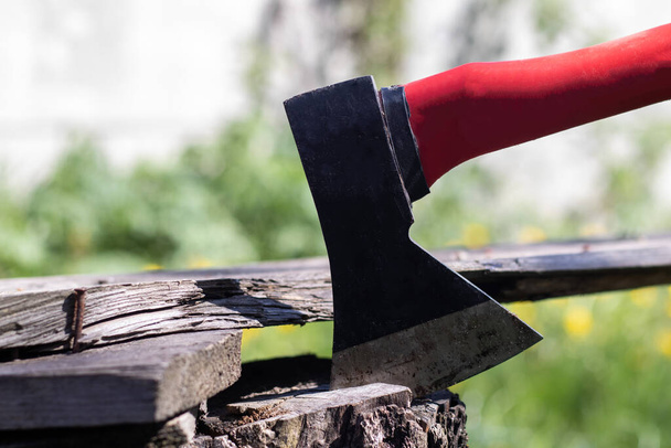 Um novo machado moderno com uma alça vermelha sai de um toco de madeira contra um prado verde em um dia ensolarado de verão. O machado ficou preso no coto. Lâmina Ax em um log - Foto, Imagem
