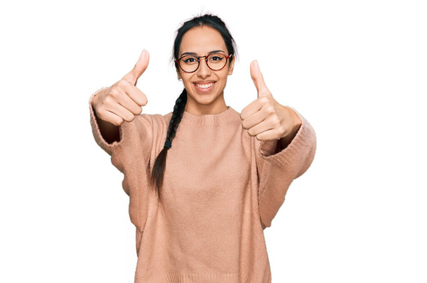 Jong Latijns-Amerikaans meisje draagt casual kleding en bril goed te keuren doen positieve gebaar met de hand, duimen omhoog glimlachend en blij voor succes. winnaar gebaar.  - Foto, afbeelding