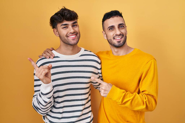 Junges hispanisches schwules Paar steht auf gelbem Hintergrund fröhlich mit einem Lächeln im Gesicht und zeigt mit der Hand und dem Finger zur Seite mit glücklichem und natürlichem Ausdruck  - Foto, Bild