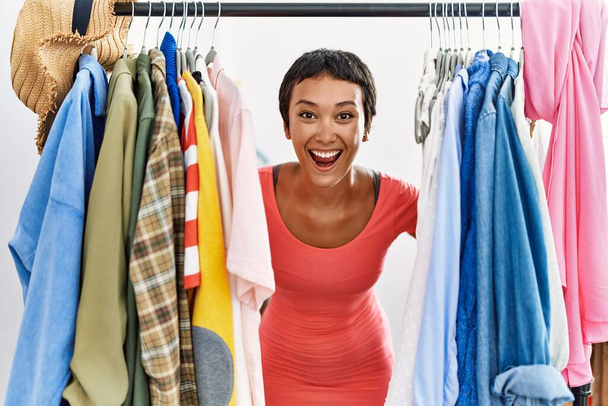 Молода іспаномовна жінка клієнт посміхається впевнено з'являється через одяг в магазині одягу
 - Фото, зображення