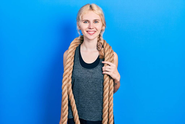 Beautiful caucasian woman with blond hair training with battle rope looking positive and happy standing and smiling with a confident smile showing teeth  - Photo, image