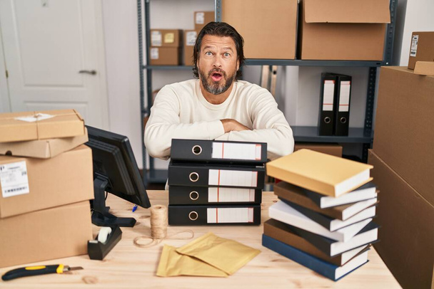 Handsome middle age man working at small business ecommerce afraid and shocked with surprise and amazed expression, fear and excited face.  - 写真・画像