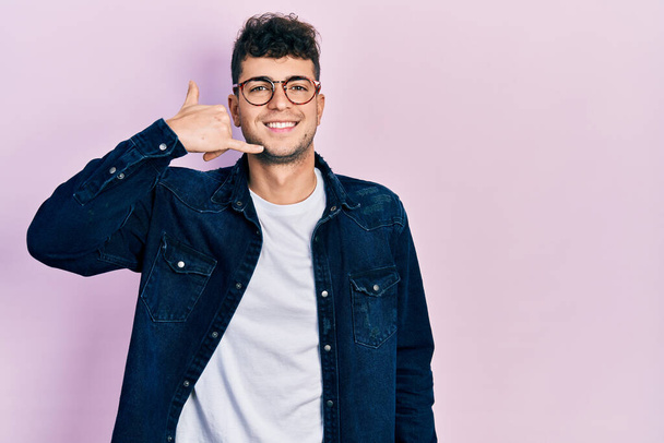 Молодий іспаномовний чоловік в повсякденному одязі і окулярах посміхається робити телефонний жест з рукою і пальцями, як розмовляти по телефону. концепції спілкування
.  - Фото, зображення