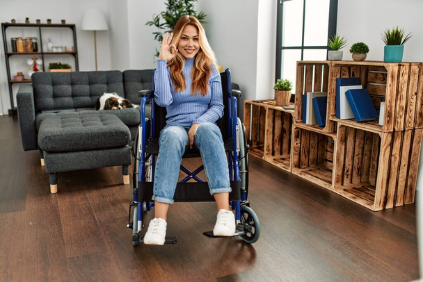 Young beautiful woman sitting on wheelchair at home smiling positive doing ok sign with hand and fingers. successful expression.  - Photo, Image