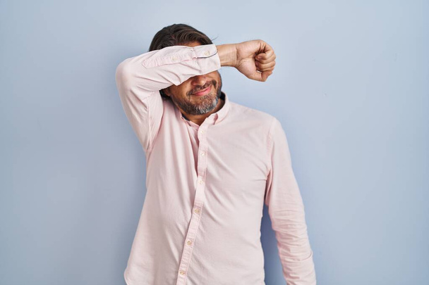 Knappe man van middelbare leeftijd draagt elegante shirt achtergrond bedekken ogen met arm lachend vrolijk en grappig. blind concept.  - Foto, afbeelding