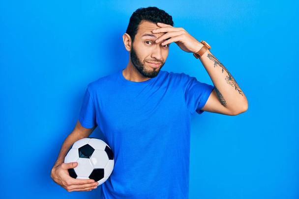 Ισπανόφωνος άνδρας με γένια κρατώντας μπάλα ποδοσφαίρου ανησυχούν και τόνισε για ένα πρόβλημα με το χέρι στο μέτωπο, νευρικός και ανήσυχος για την κρίση  - Φωτογραφία, εικόνα