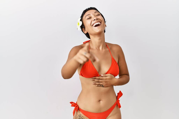 Junge hispanische Frau mit kurzen Haaren im Bikini, die dich anlacht, mit der Hand über dem Körper in die Kamera zeigt, beschämender Ausdruck  - Foto, Bild