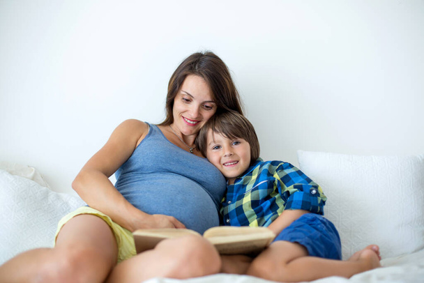 Young pregnant mother, lying in bed, reading a book to her older child, boy, daytime - Photo, Image