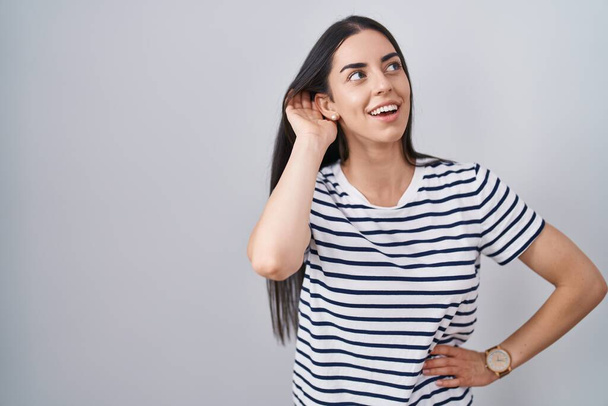 Junge brünette Frau in gestreiftem T-Shirt, die lächelnd mit der Hand über dem Ohr Gerüchten oder Klatsch lauscht. Konzept für Gehörlosigkeit.  - Foto, Bild