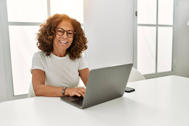 Middelbare leeftijd Spaanse vrouw glimlachen zelfverzekerd met behulp van laptop thuis - Foto, afbeelding