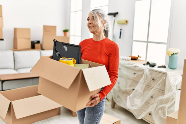 Mujer de pelo gris de mediana edad sonriendo confiado sosteniendo paquete en el nuevo hogar - Foto, imagen