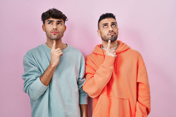 Молода пара іспаномовних геїв, що стоять над рожевим фоном, думає про сумніви з пальцем на підборідді і дивлячись дивуючись
  - Фото, зображення
