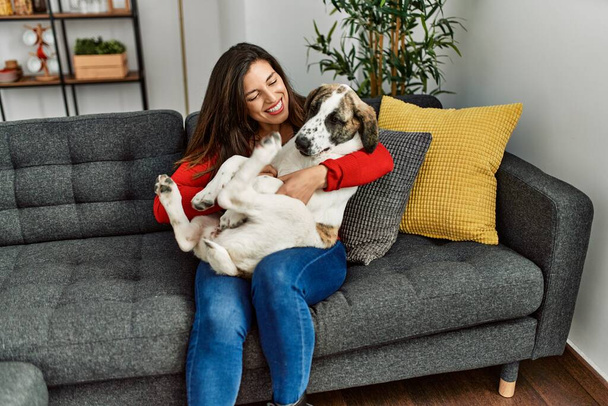 Νεαρή γυναίκα αγκαλιάζει το σκυλί κάθεται στον καναπέ στο σπίτι - Φωτογραφία, εικόνα