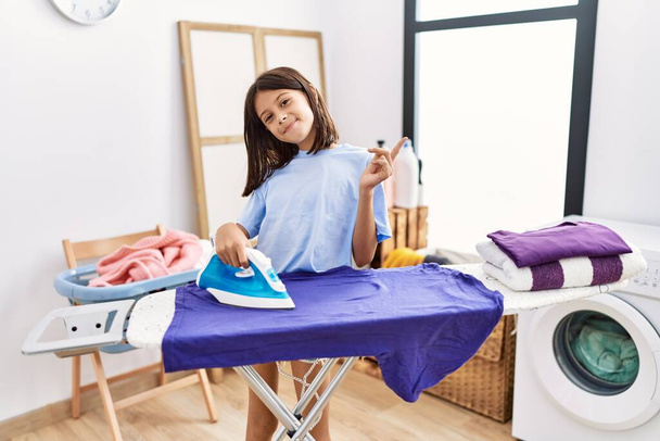 Young hispanic girl ironing clothes at laundry room smiling happy pointing with hand and finger to the side  - Photo, Image