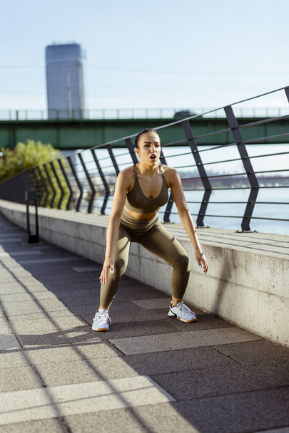 Pretty young woman in sportswear stretching on a river promenade - Foto, afbeelding