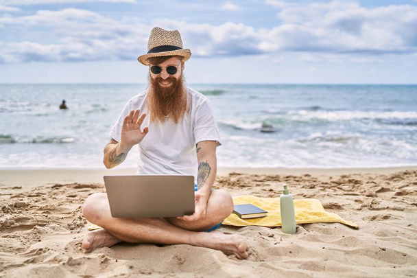 Молодий рудий чоловік має відеодзвінок за допомогою ноутбука на пляжі
. - Фото, зображення