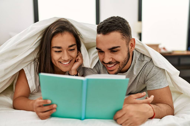 Fiatal latin pár olvasás könyv borító ágynemű az ágyon. - Fotó, kép