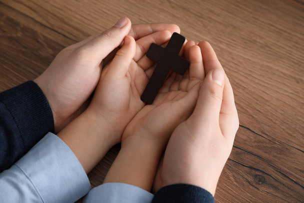 Boy and his godparent holding cross at wooden table, closeup - Foto, Bild