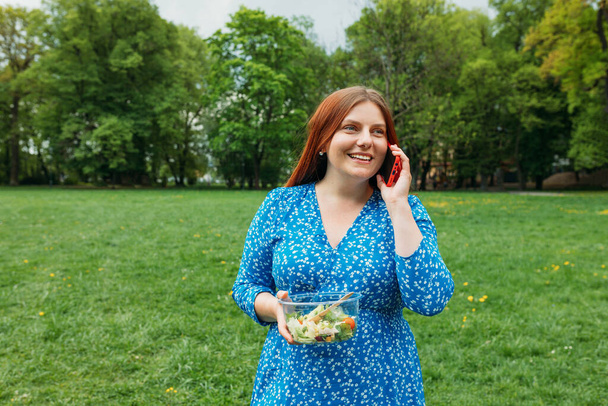 Un retrato de una hermosa mujer sonriente hablando por teléfono sobre el fondo de la naturaleza. Mujer feliz sosteniendo tazón con ensalada y usando un teléfono inteligente en el parque al aire libre - Foto, Imagen