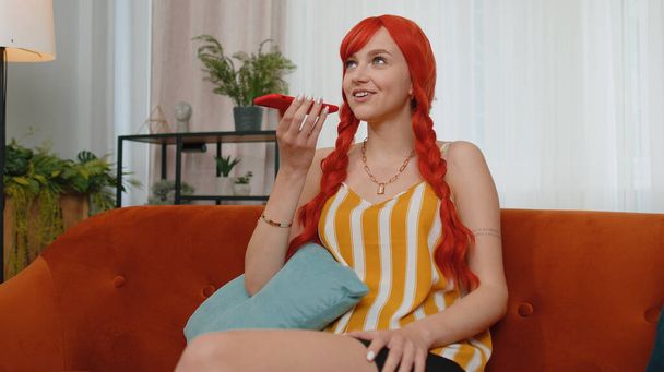 Улыбающаяся рыжая девушка в майке-цистерне разговаривает по телефону с друзьями, сидящими на диване в комнате. Счастливая возбужденная рыжая деловая девушка наслаждается мобильный громкоговоритель говорить в помещении - Фото, изображение