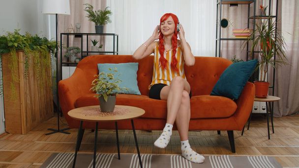 Happy relaxed overjoyed redhead girl in wireless headphones dancing on couch at home choosing listening favorite energetic disco rock n roll music in smartphone entertaining. People weekend activities - Photo, Image