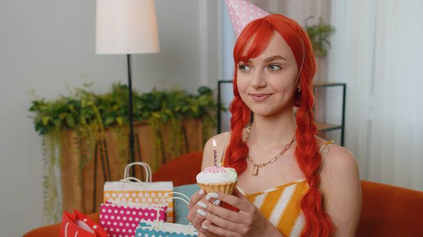 お祝いの誕生日の帽子の幸せな赤い髪の女の子はカップケーキを保持しているケーキの上に燃えるろうそくを吹くことを喜んでお祝いします。若いです生姜女性お祝いパーティーでソファにギフトで家 - 写真・画像