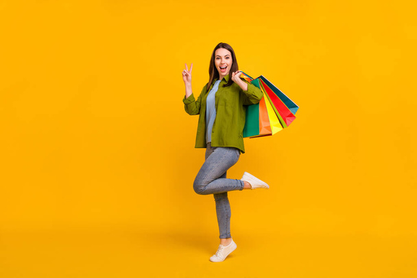 Photo of sweet excited young woman dressed green shirt holding bargains showing v-sign smiling isolated yellow color background. - Fotoğraf, Görsel