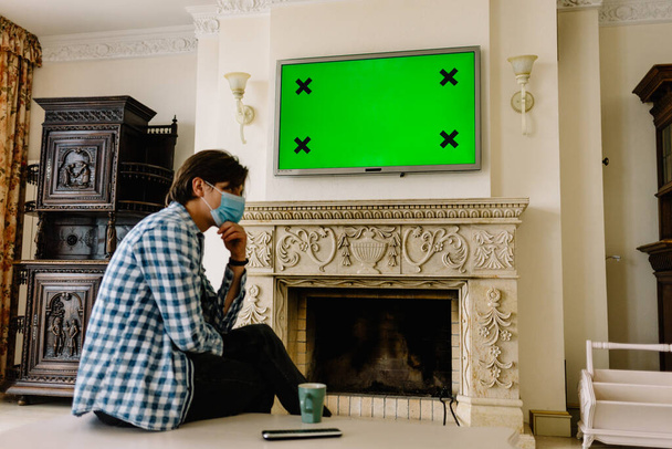 El joven con una máscara médica mira la pantalla verde de la televisión desde casa, está en cuarentena de la epidemia de coronavirus, covid-19. Quiere trabajar y está esperando a que aparezca la vacuna contra el virus. Nueva realidad - Foto, Imagen